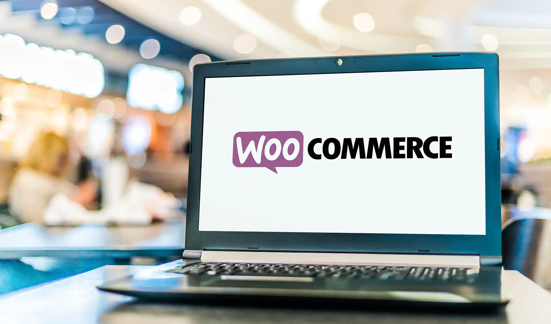 A laptop on a table displays the WooCommerce logo on its screen, while blurred lights and people fill the background—perfectly capturing how Sequential Order Numbers for WooCommerce streamlines sales tracking in any busy indoor setting.