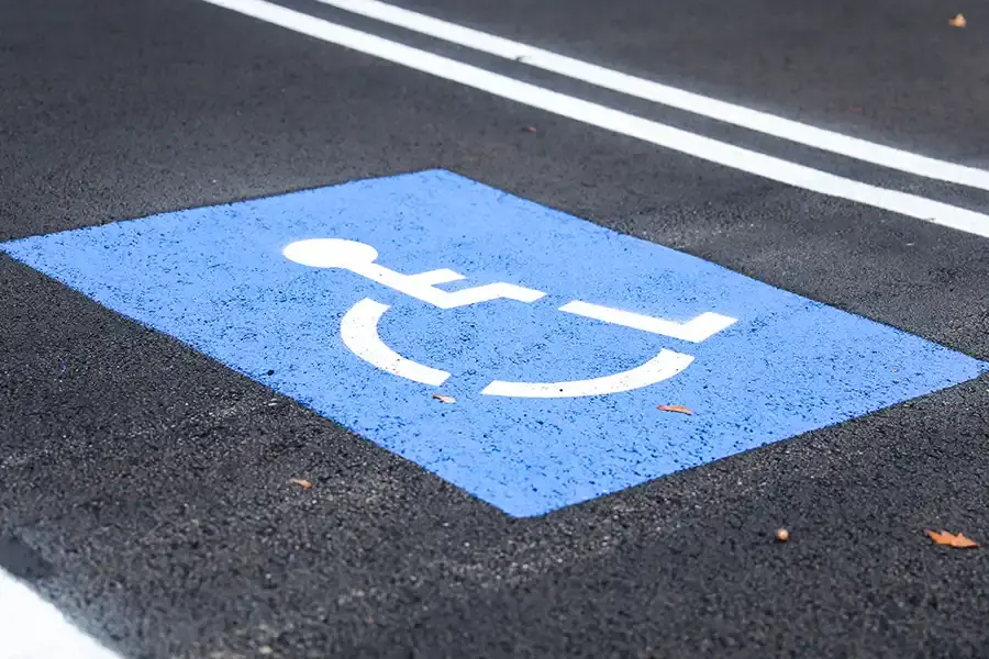 A blue parking spot with a white wheelchair symbol painted on asphalt, indicating an ADA-compliant accessible parking space. Two white parallel lines are visible in the background.