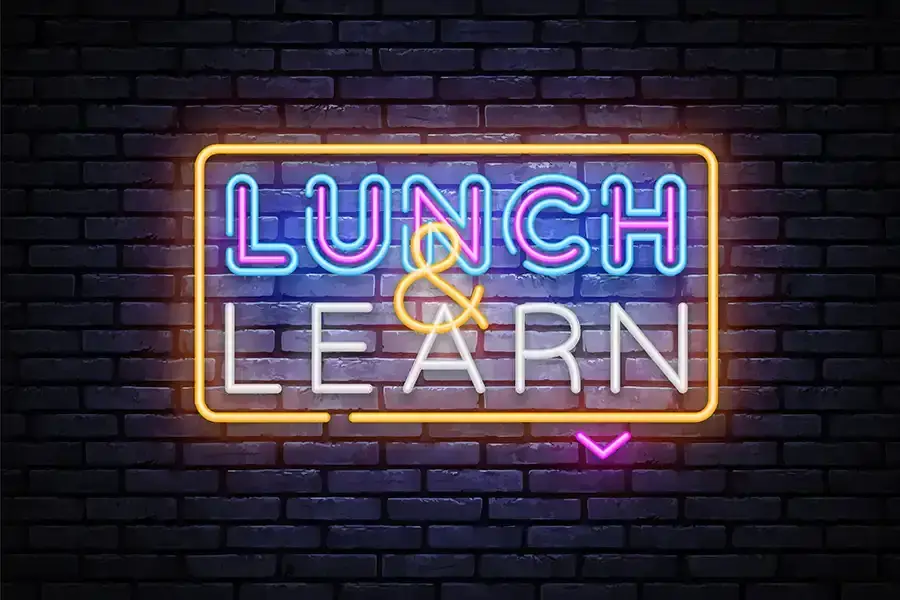A neon sign on a dark brick wall reads Lunch and Learn in bright blue, pink, yellow, and white colors, glowing with a vivid outline and a small pink arrow pointing below the word LEARN.