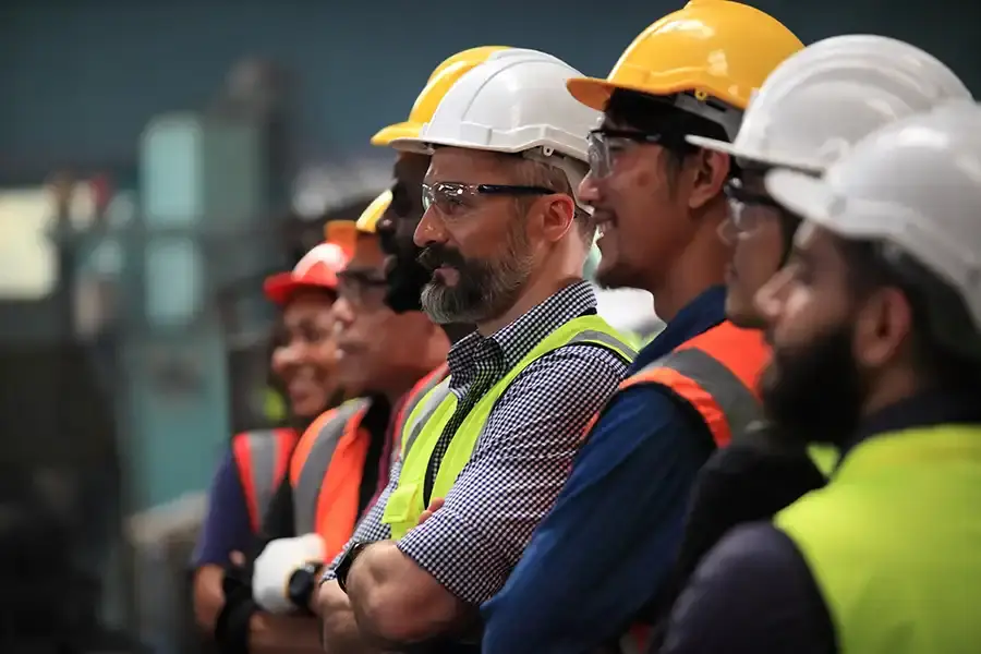 A group of workers wearing safety helmets, vests, and protective eyewear stand side by side at an industrial worksite, confident despite concerns that Federal Contract Reforms Are Shutting Out Small Businesses.